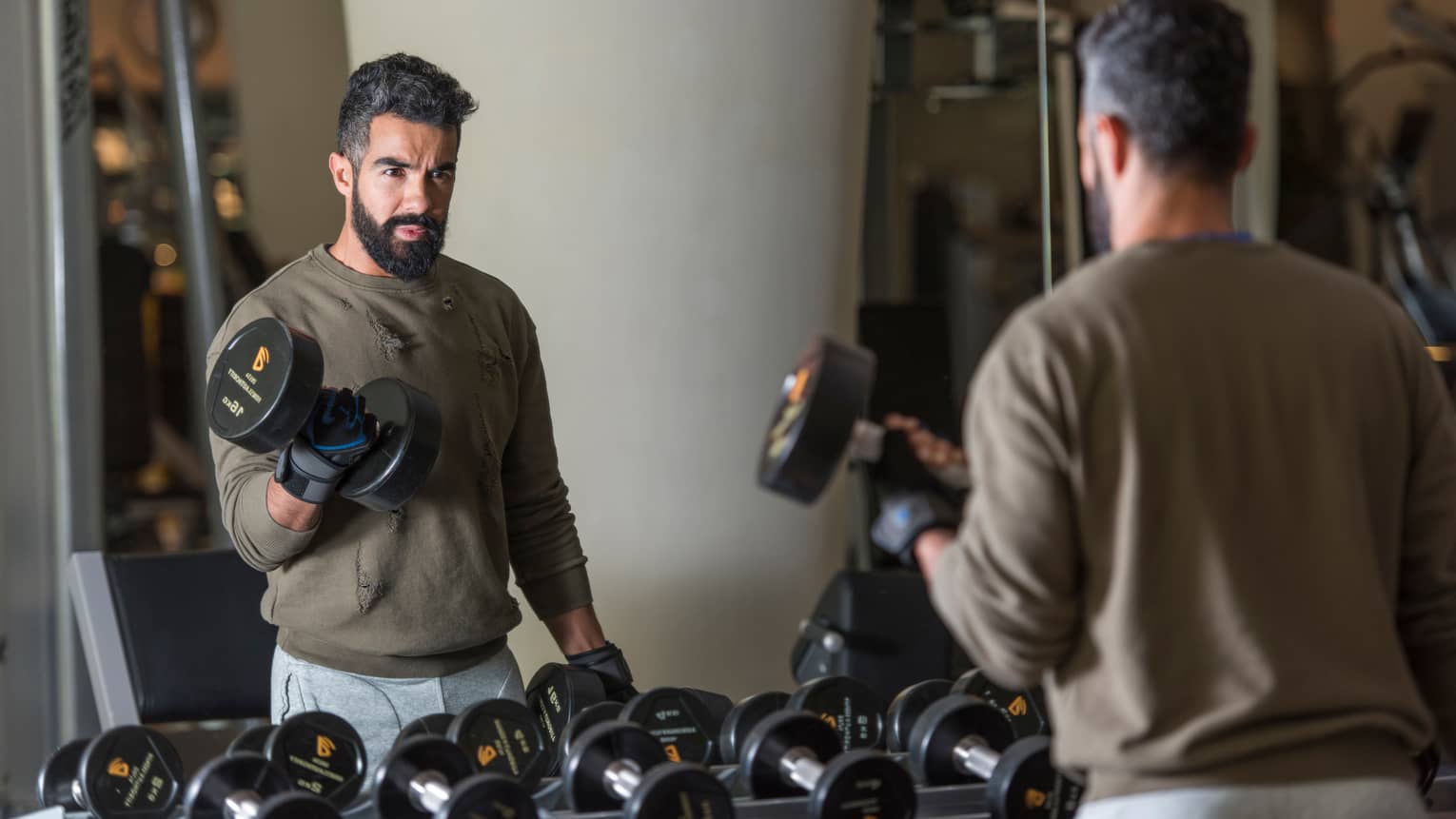 Man with beard wearing casual sweatshirt and sweatpants stands looks in gym mirror, lifts large hand weights