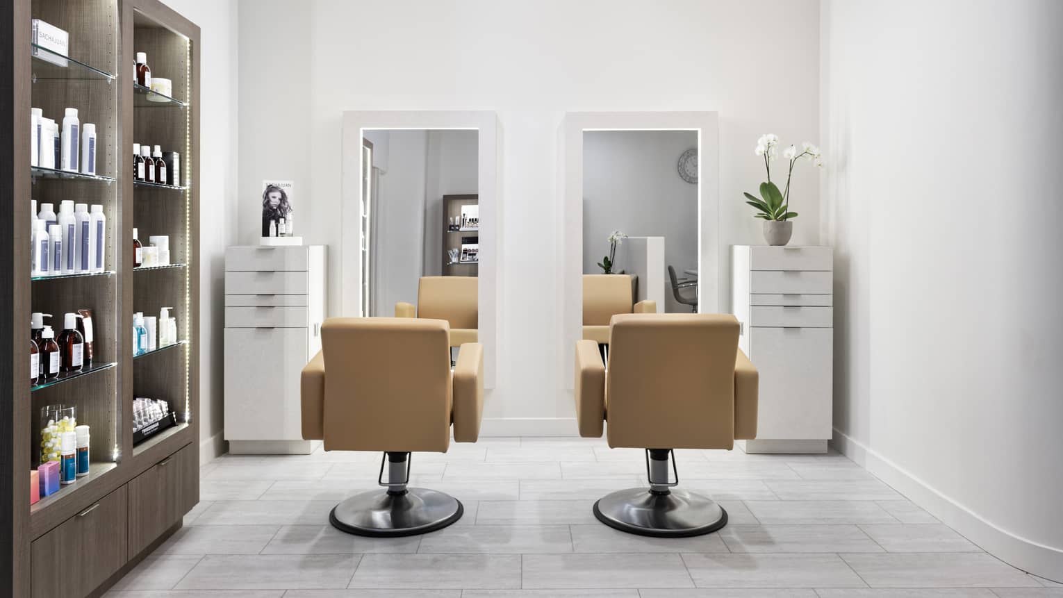 A hair salon with two light brown leather chairs facing mirrors, wall of products