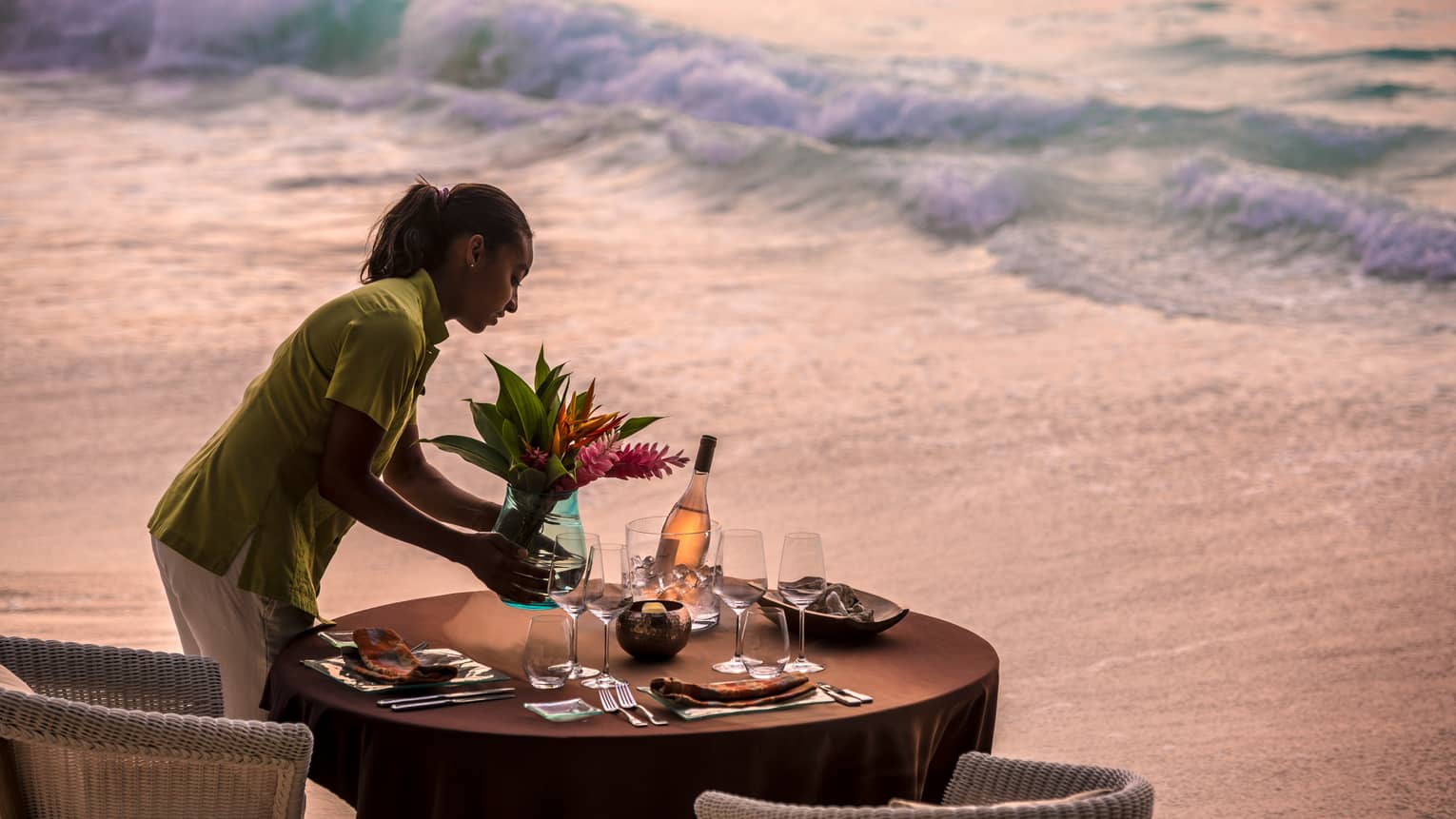 Woman sets vase with flowers on round dining table in front of beach tides at sunset 