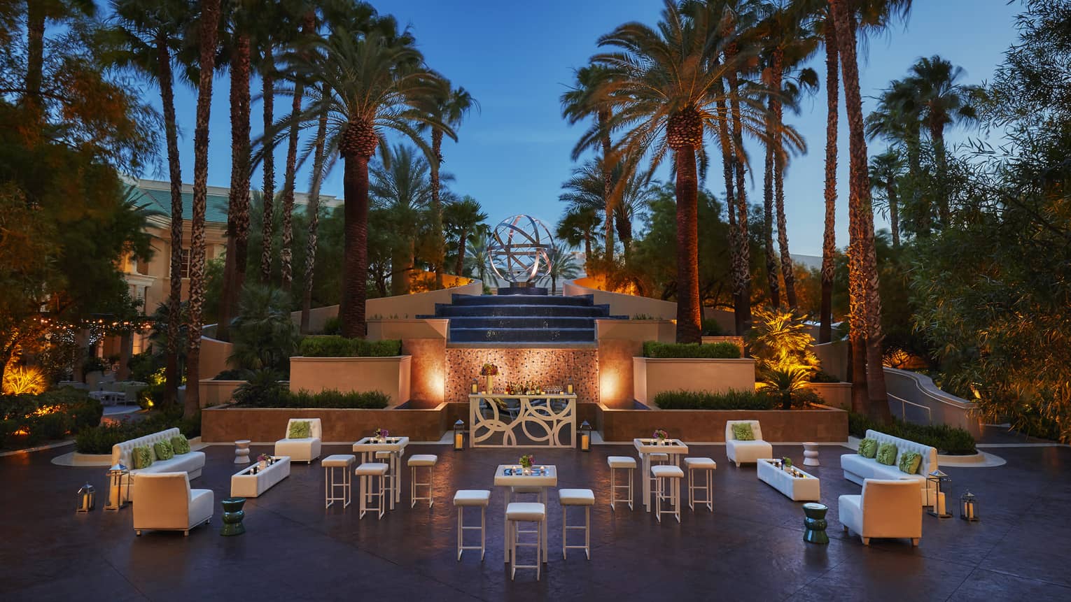 Palm trees over stone fountain, terrace seating area with lights at dusk