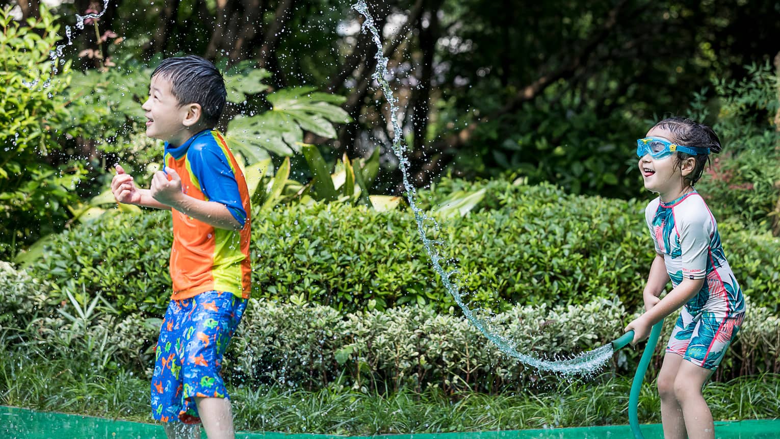 Young girl wearing swimsuit, goggles sprays boy with garden hose