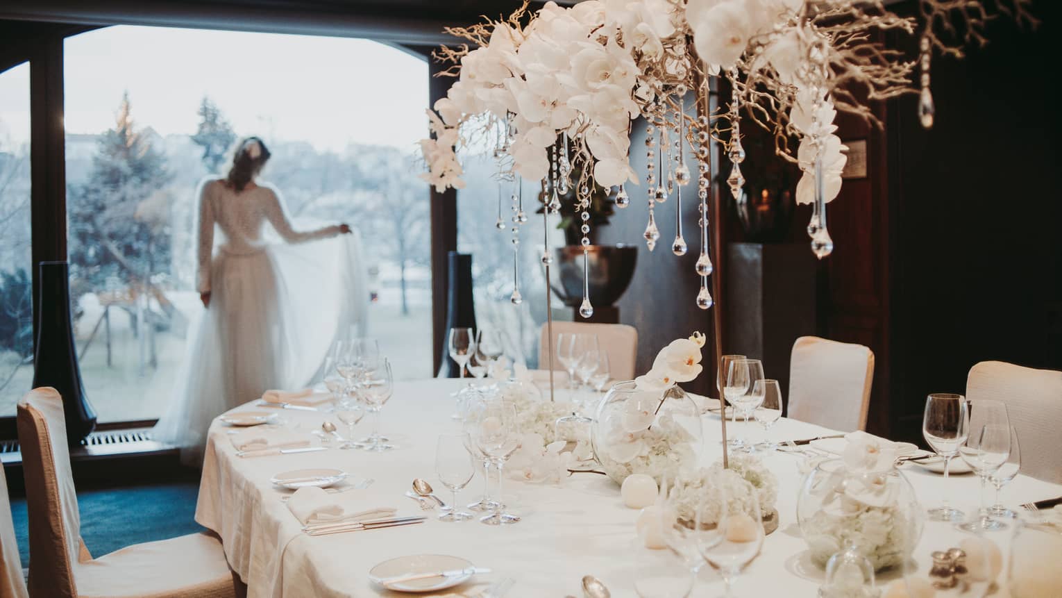Bride holds train staring out window behind table with white orchids and glass icicles 