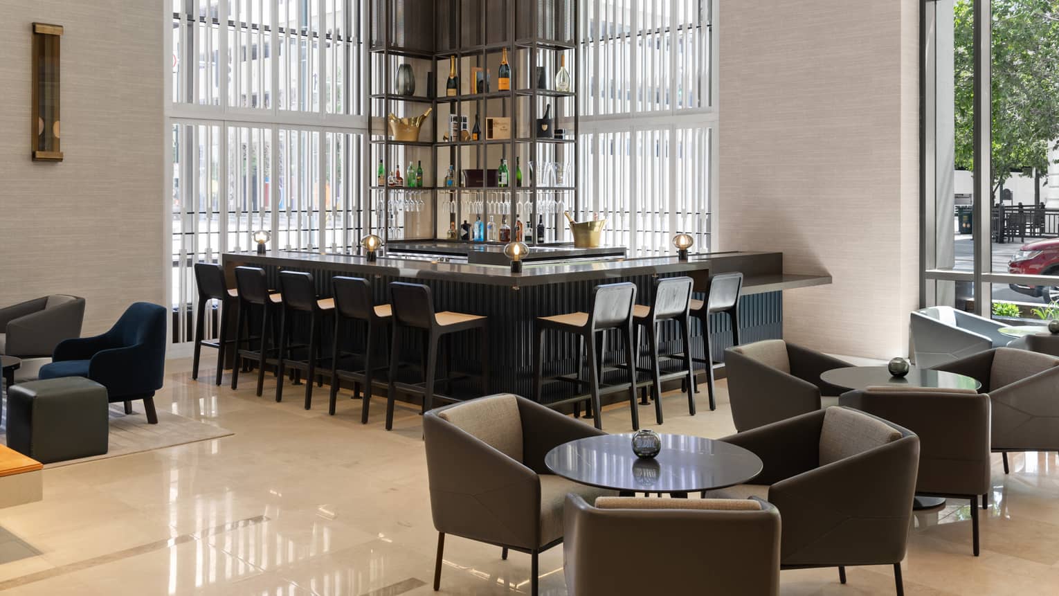 A black bar and lounge by large windows surrounded by black and wood stools and round tables with grey chairs.