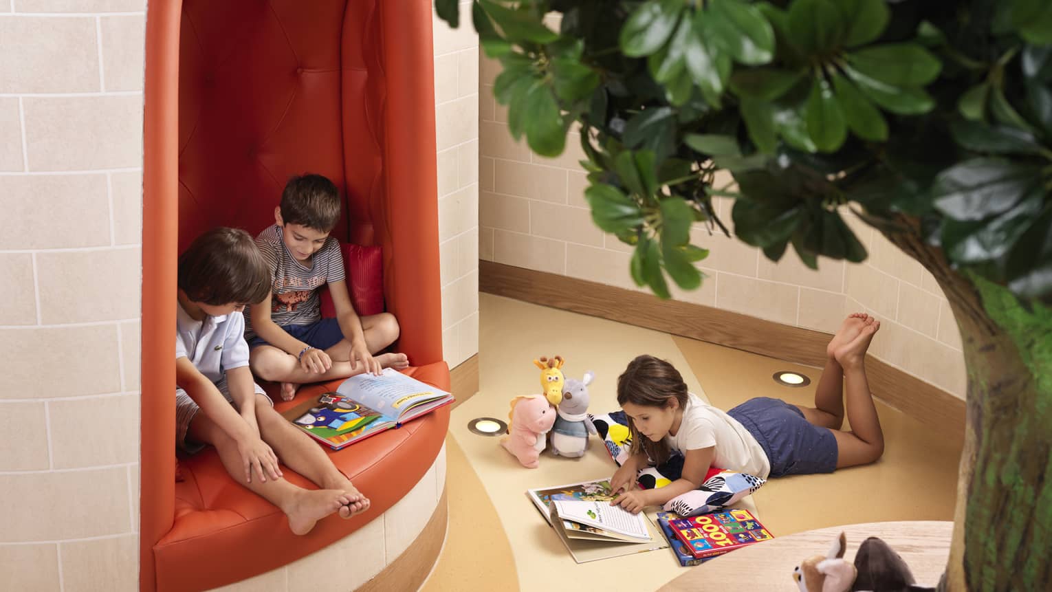 A child lays on the floor reading a book while two other children sit in a red tufted booth reading another book