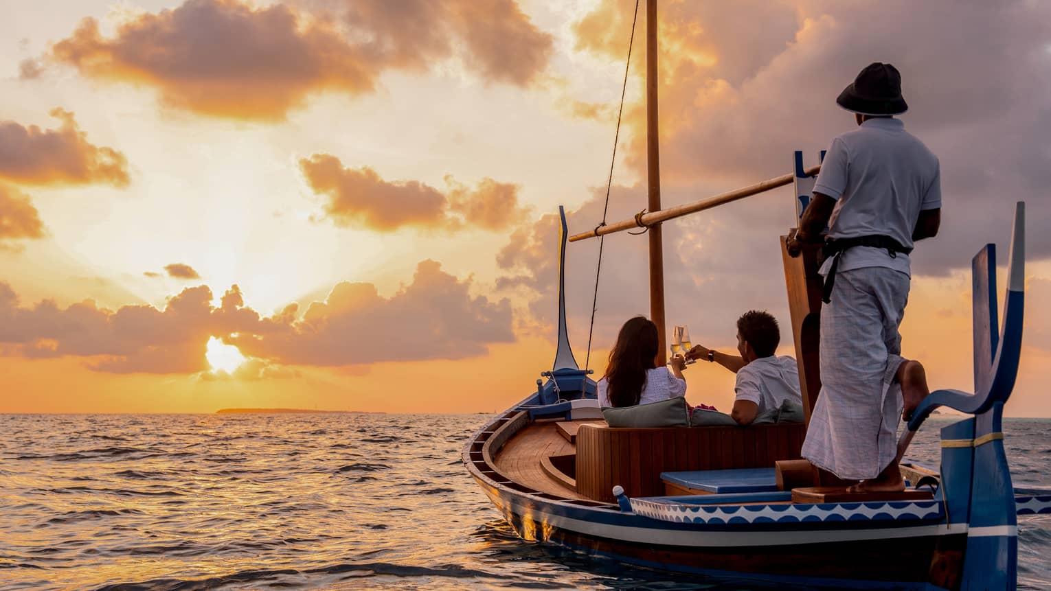 Rear view of two people seated in a dhoni boat, toasting with champagne, the boat handler steering into the golden sunset.