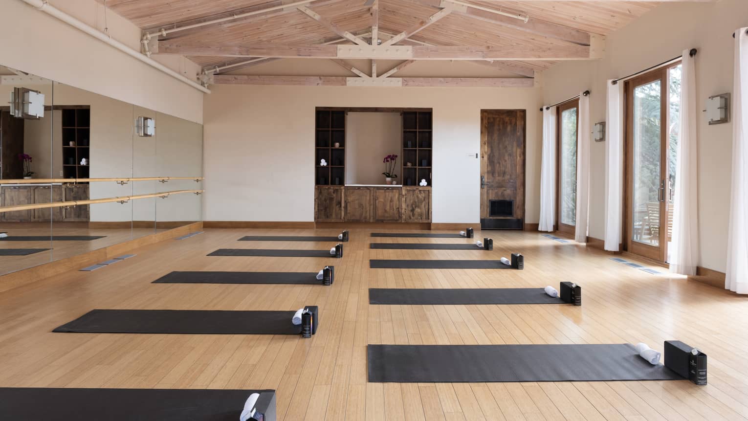 Interior of yoga studio with open rafter ceilings, one mirrored wall and wood floors with two rows of black yoga mats set out on the floor
