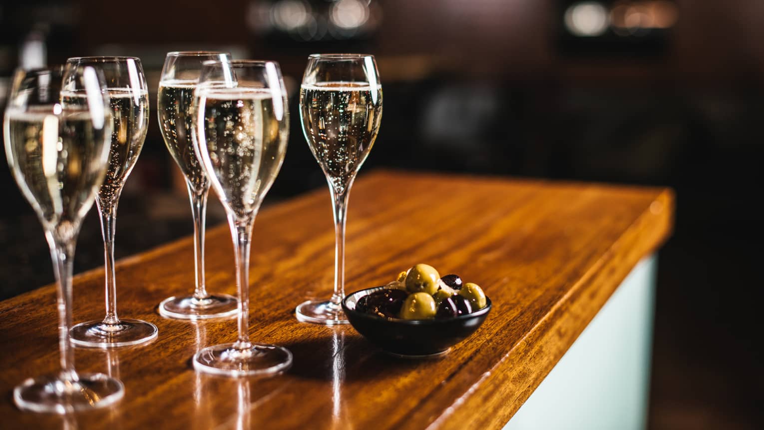 Five glasses of sparkling wine with bowl of black and green olives, atop a wooden bar top