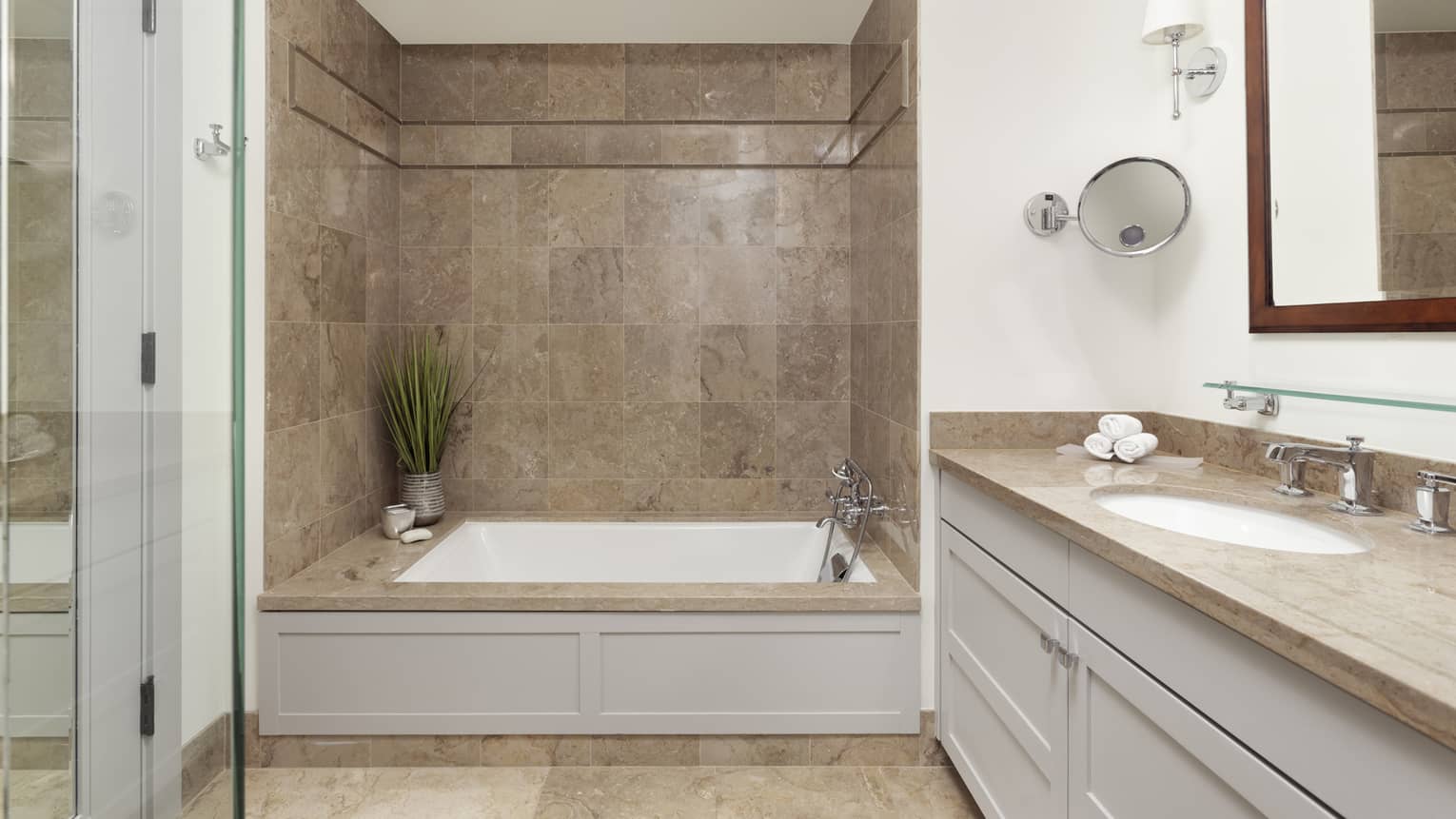 A private residence bathroom has a tan stone shower with a white tub, white cabinets with tan stone counter 