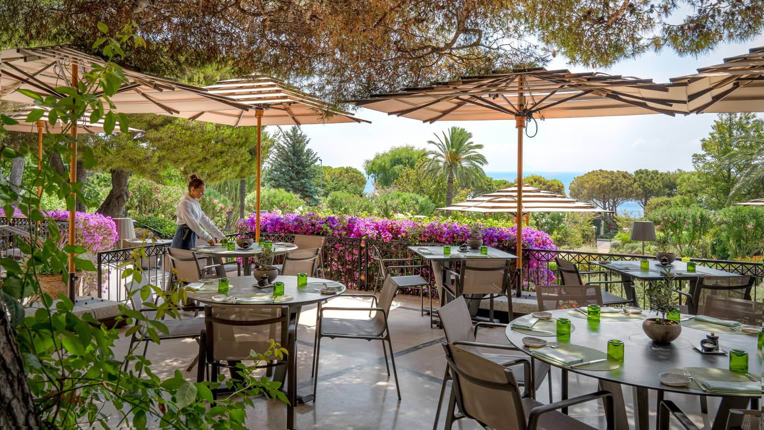 Waiter sets a table on outdoor restaurant terrace, surrounded by greenery and magenta-coloured flowers