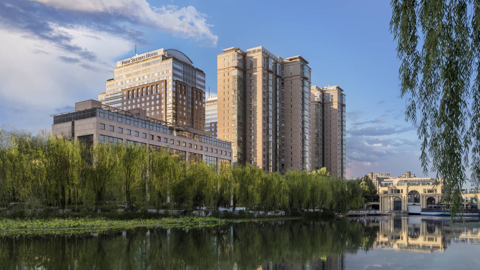 A large hotel building on the edge of a body of water, a line of tall green plants grows near the water.