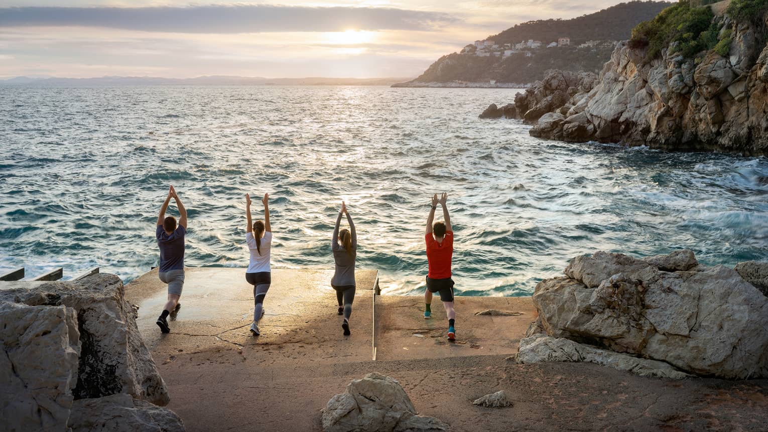 Rear view of four people doing yoga on sandy shore next to sea, rocky cliffs on right