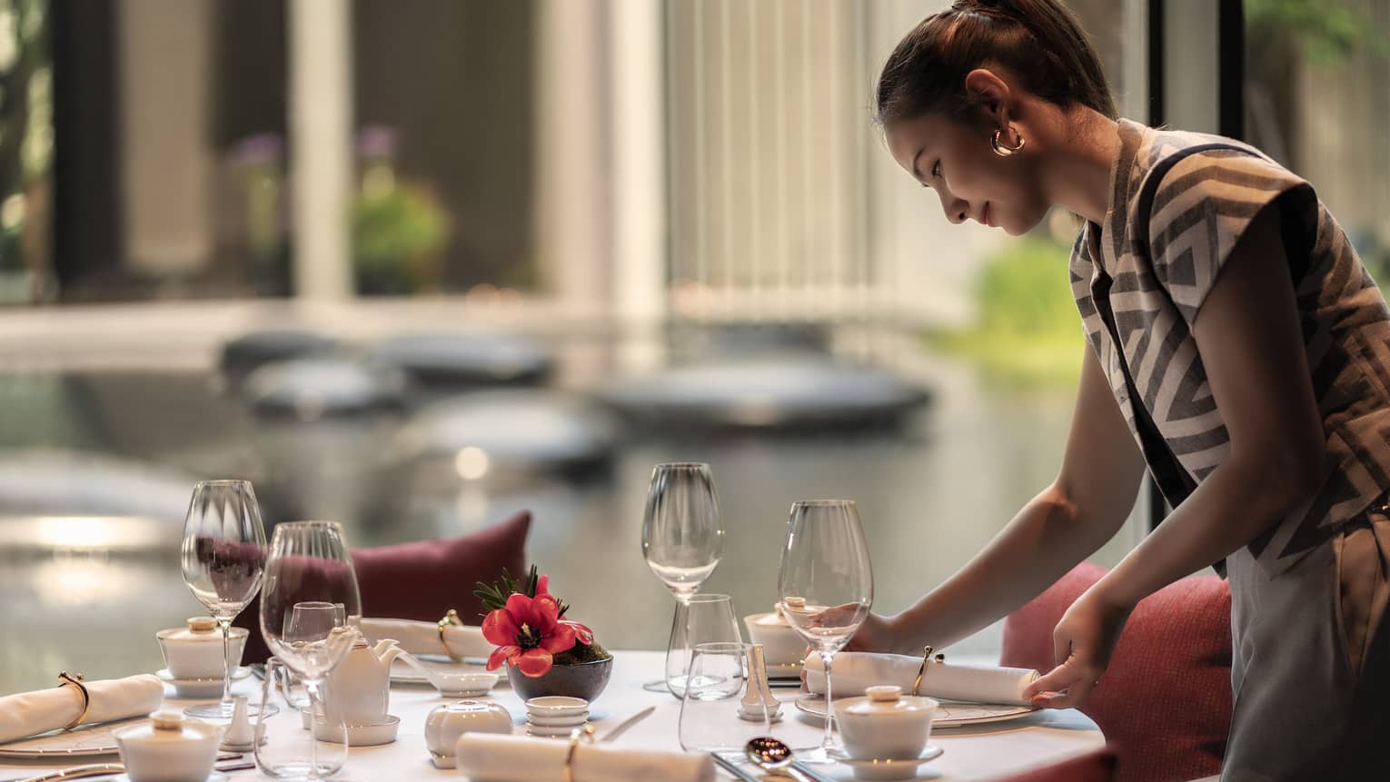 Woman sets a round table at restaurant, looking out to a tranquil pond