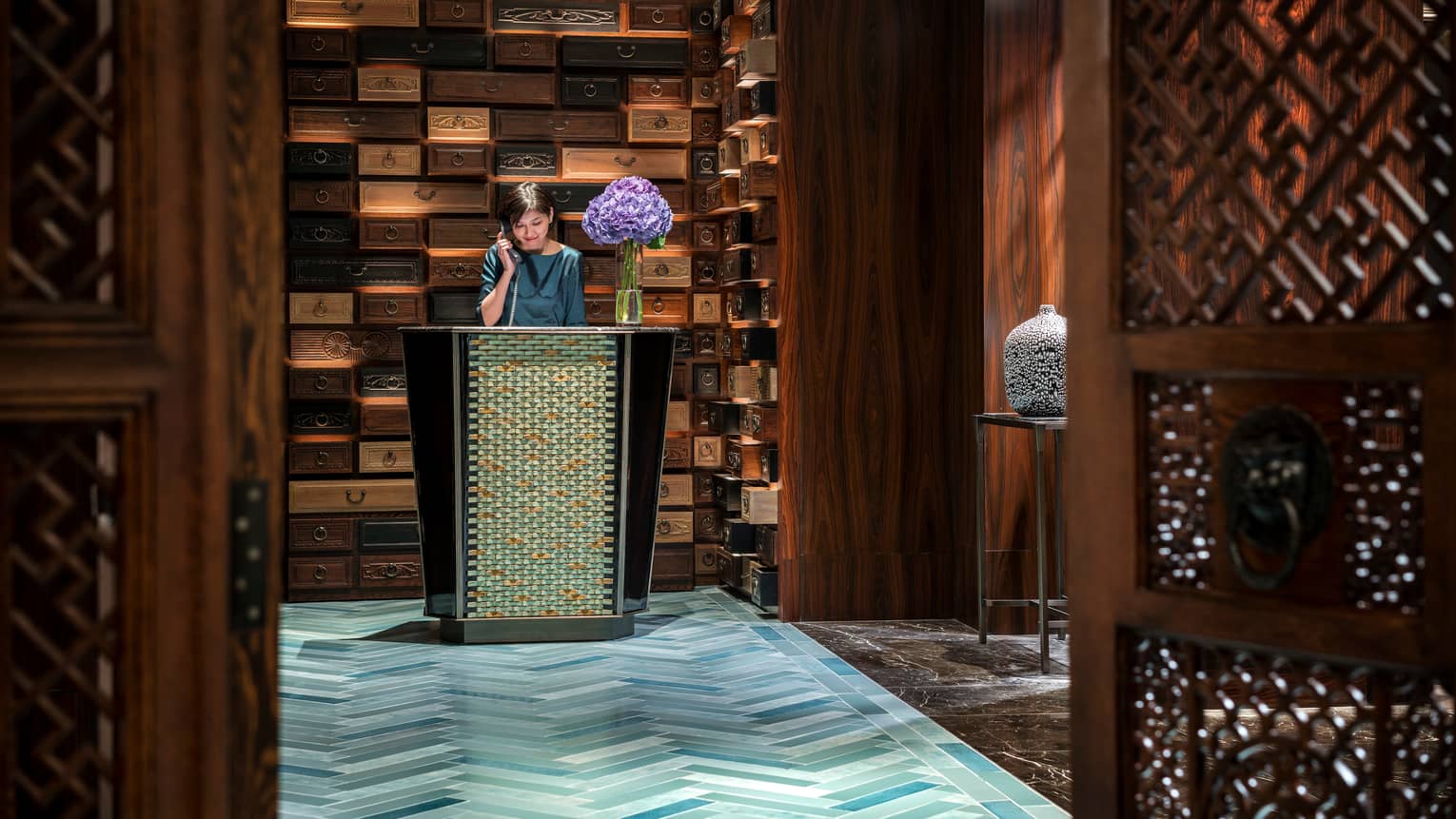 Hotel staff answers telephone at reception desk in front of decorative wall made from wood drawers