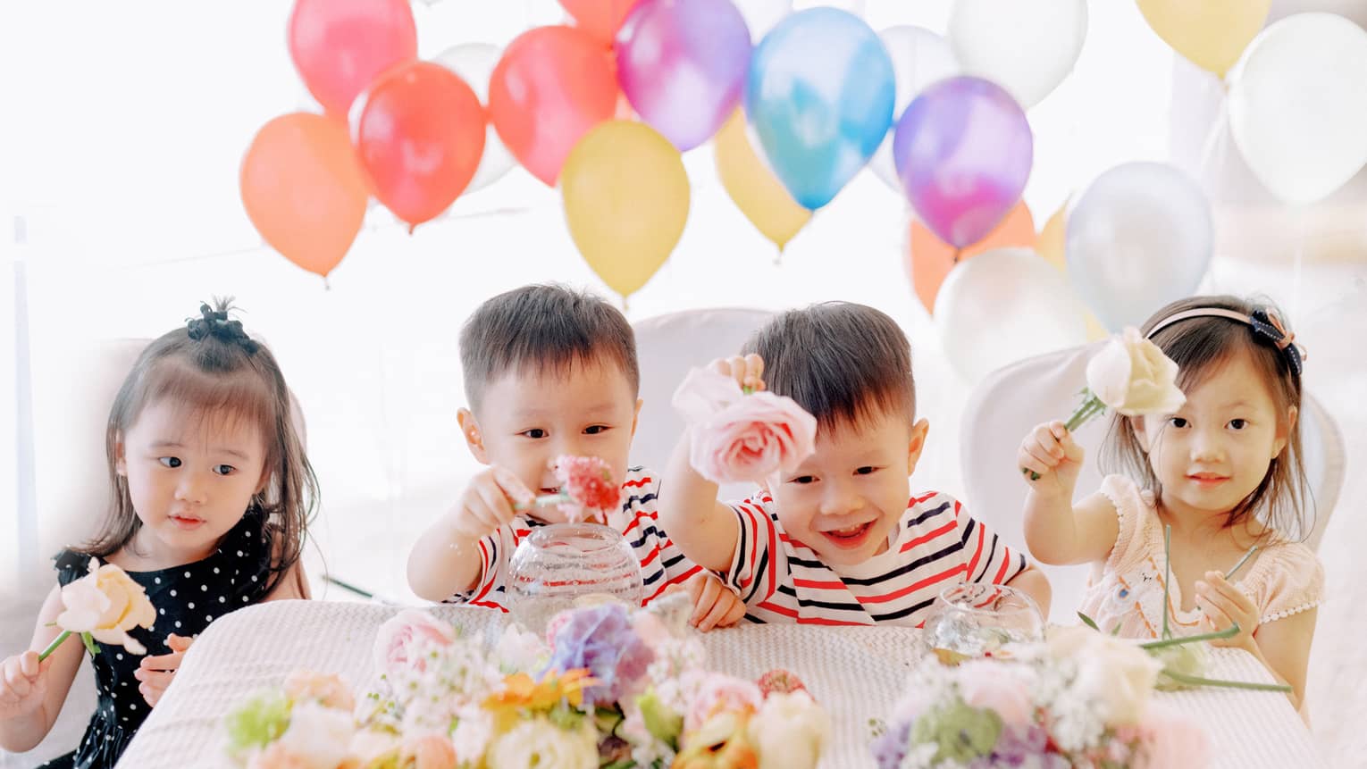 Four toddler children holding roses at table with balloons behind them