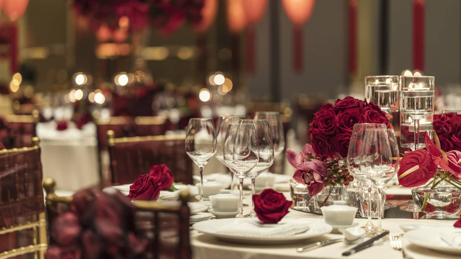 Elegant wedding banquet table with red roses, glassware, hanging paper lanterns in background