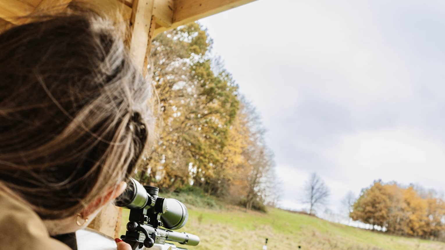 Rear view of a person in a brown jacket aiming an air rifle at small, distant targets in a large field amid autumn trees.