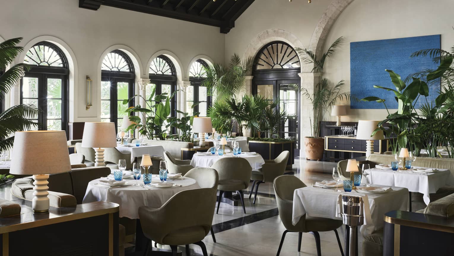 The inside of a restaurant with white linen cloth on tables, a large chandelier and beige walls.