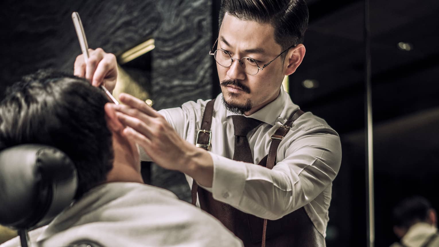 Barber wearing tie, apron combs man's hair as he sits in barber's chair