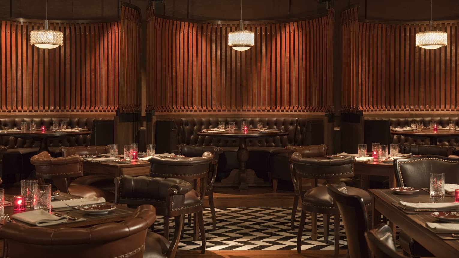 Dimly lit lounge with tufted dark brown leather booths, tables with wood-and-leather chairs, a black-and-white parquet floor and small red candles on every table