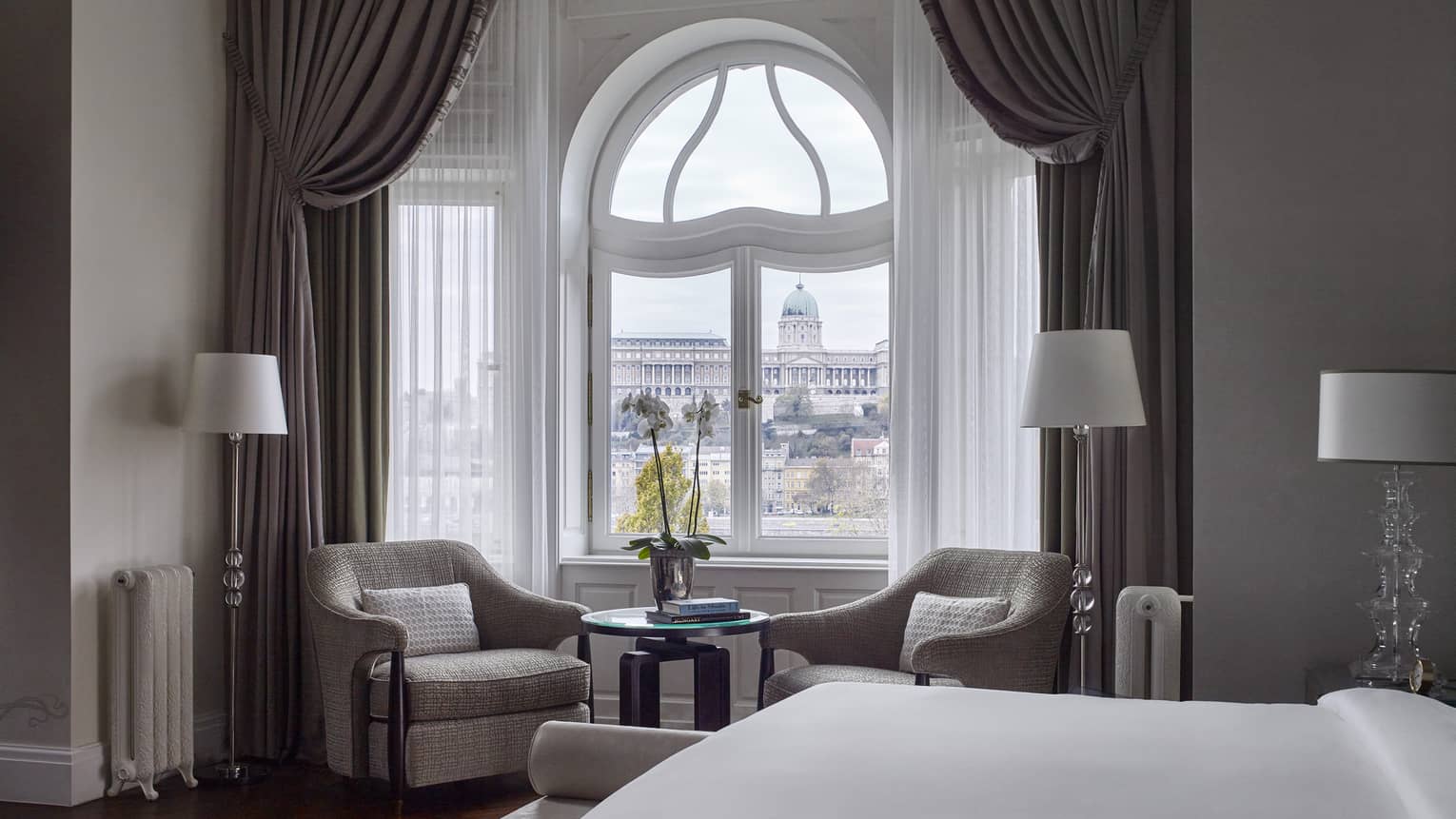 Buda Castle Presidential Suite is furnished with a two sitting chairs next to a curved window, lamps, a large bed and gray curtains. 