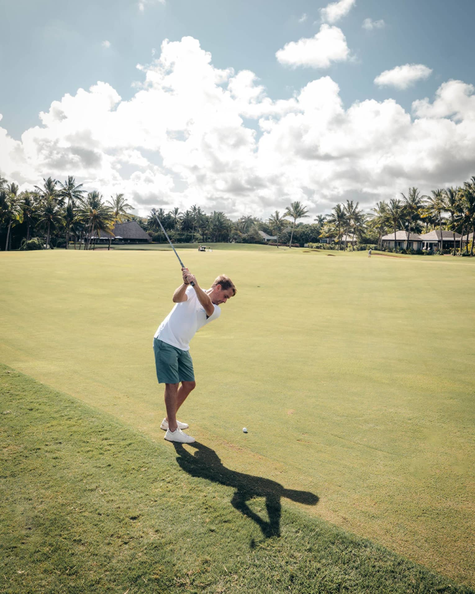 Man in white polo and green shorts swinging a golf club on green with palm trees and cloud-dotted sky in backdrop