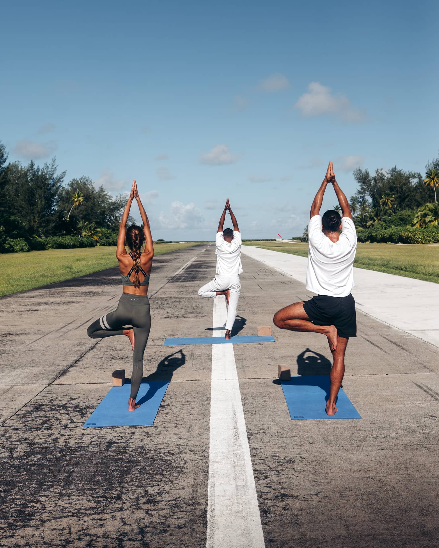 Rear view of three people practicing yoga in tree pose on an airport runway framed by lush greenery, under a clear blue sky.