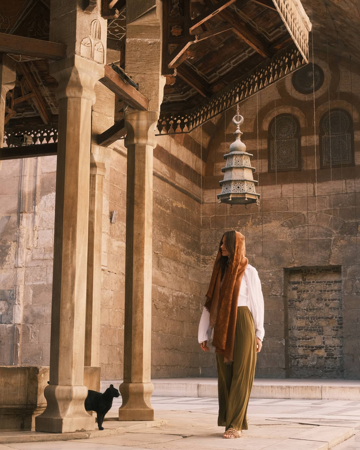 A person, head covered, stands under a pyramidal mosque lantern amid ornately carved walls and columns, a black cat nearby.