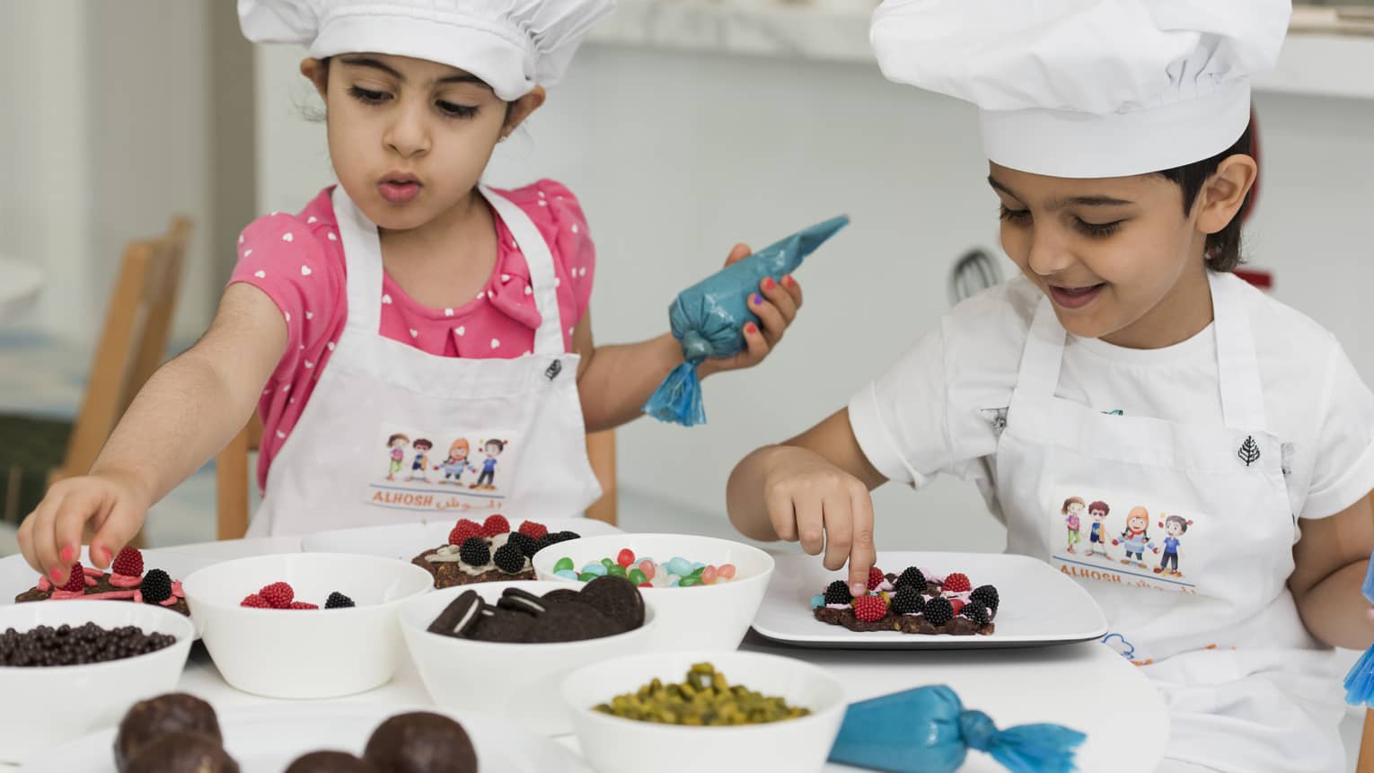 Two young girls in chef hats and aprons decorating cookies.