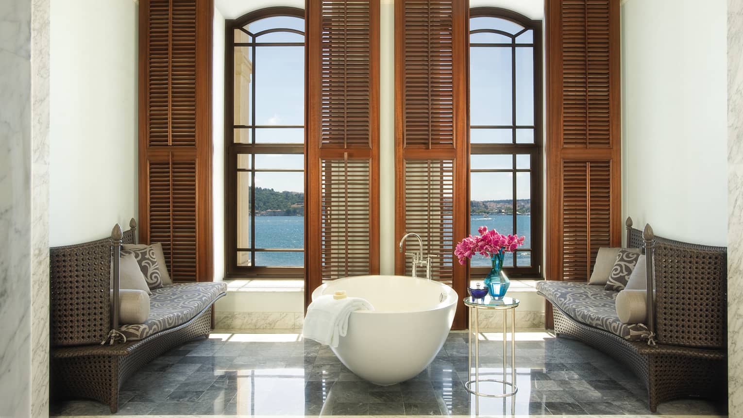 White freestanding tub in front of tall windows, wood shutters in marble bathroom