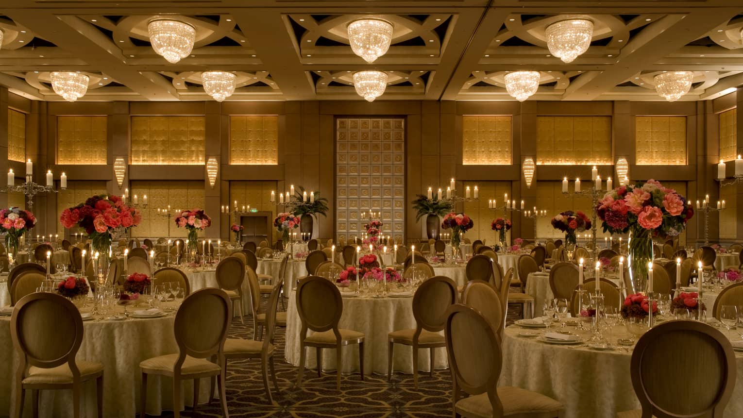 Dimly lit event room with chandeliers, tables set with large floral displays, glasses and candles 