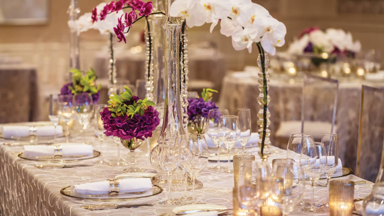 A fancy long table setting with white orchids and other purple flowers and candles as the centerpiece