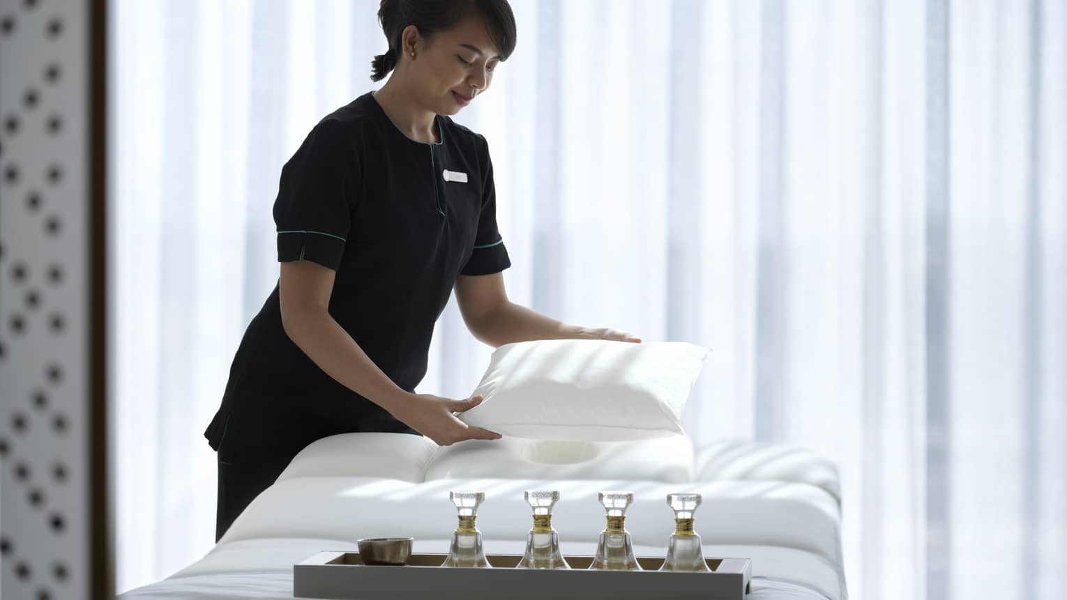 Spa attendant wearing a black uniform fluffs a while pillow on a treatment table covered with all-white linens and a tray filled with four small glass bottles