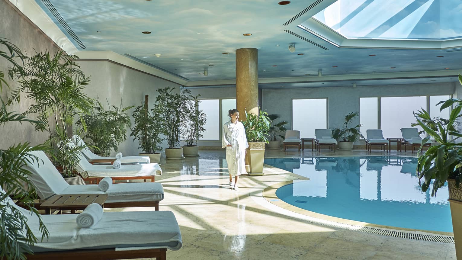 Woman in white bathroom walks the length of the indoor pool with skylight; potted plants and pool longues surrounding