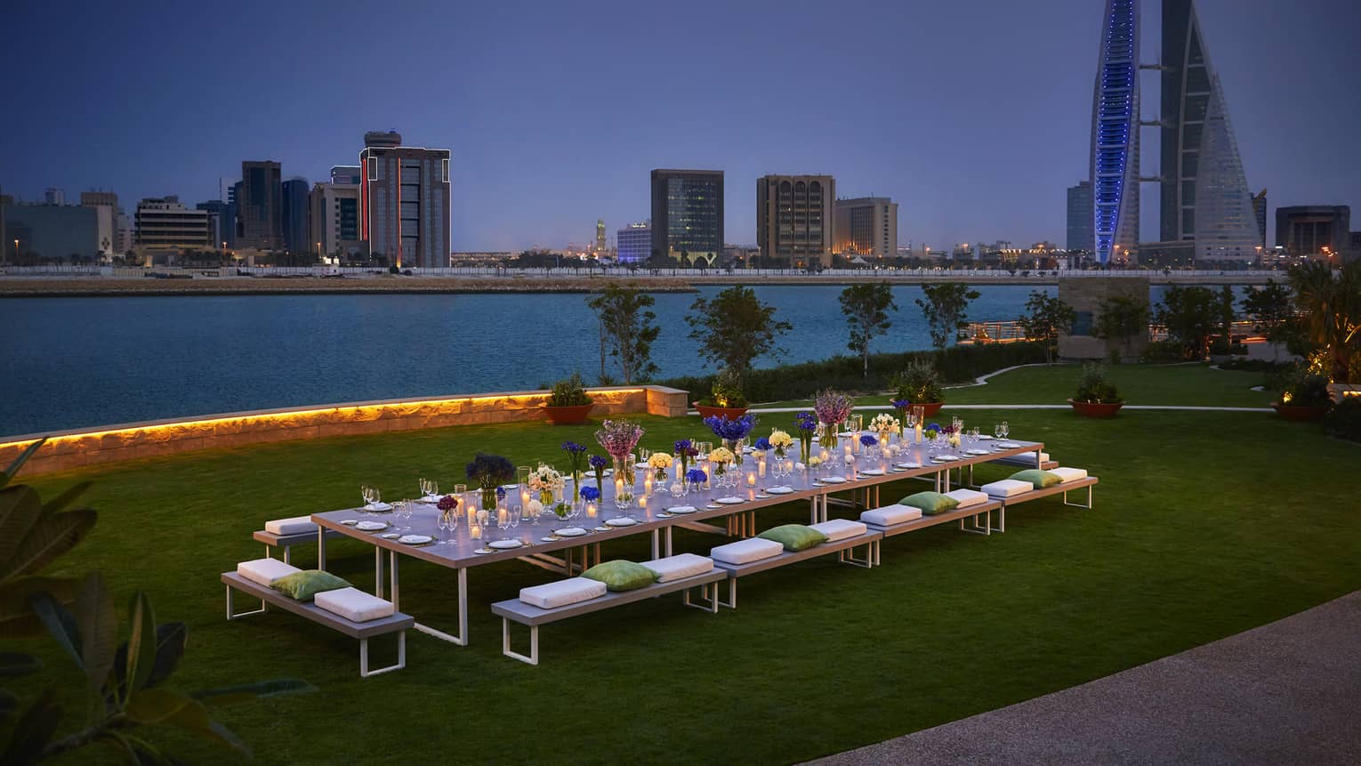 Long, candle-lit dining table on event lawn by water at night