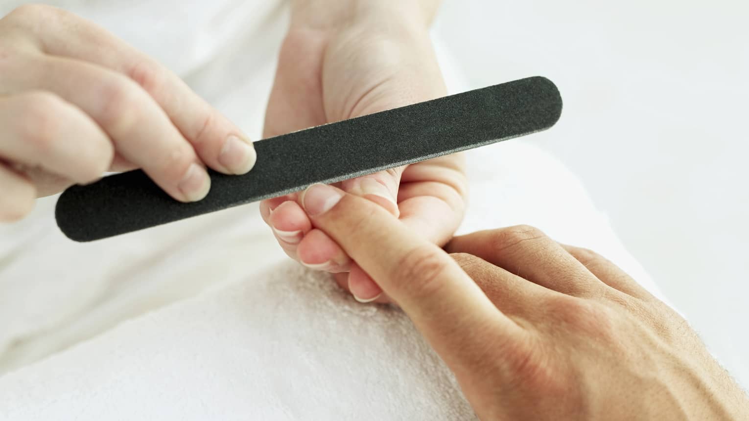 Woman holds manicure nail file to man's fingernails over white towel