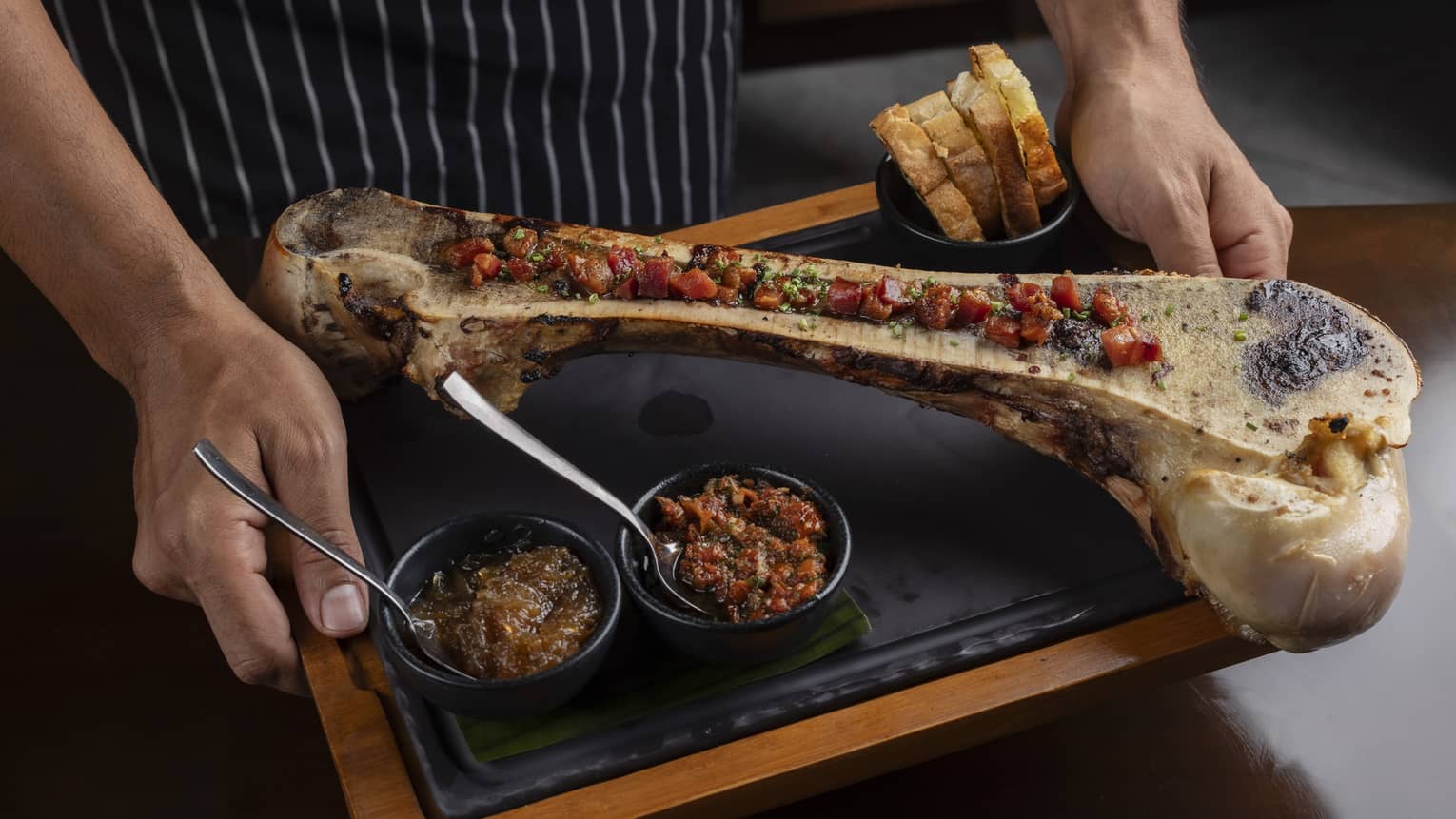 Large piece of bone cut in half and topped with prosciutto, chimichurri, cives and chiverre jam, served with a side of toasted bread all on a wood-and-black serving platter