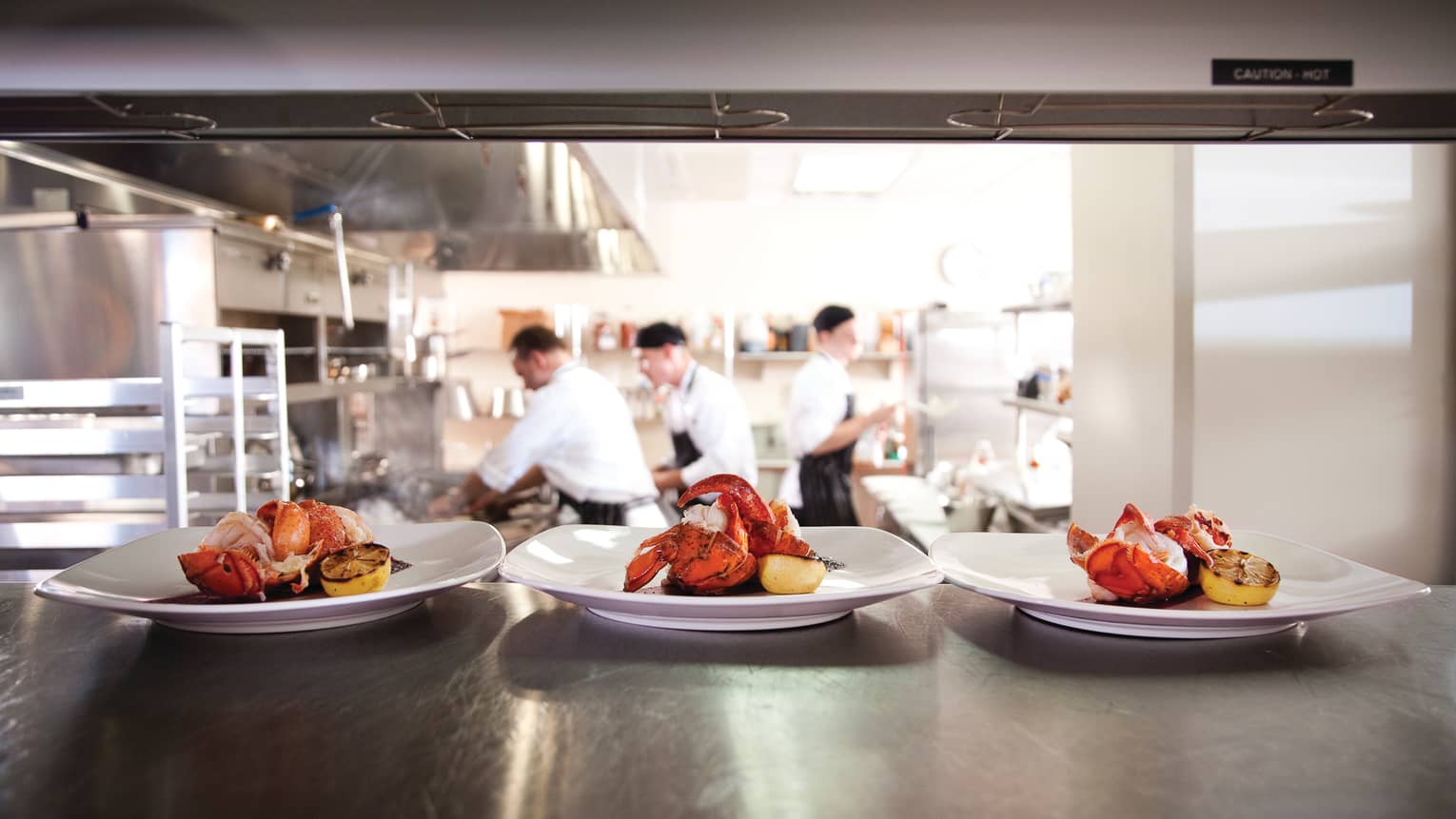 Three white plates with whole lobsters, lemons on kitchen lines, chefs in background