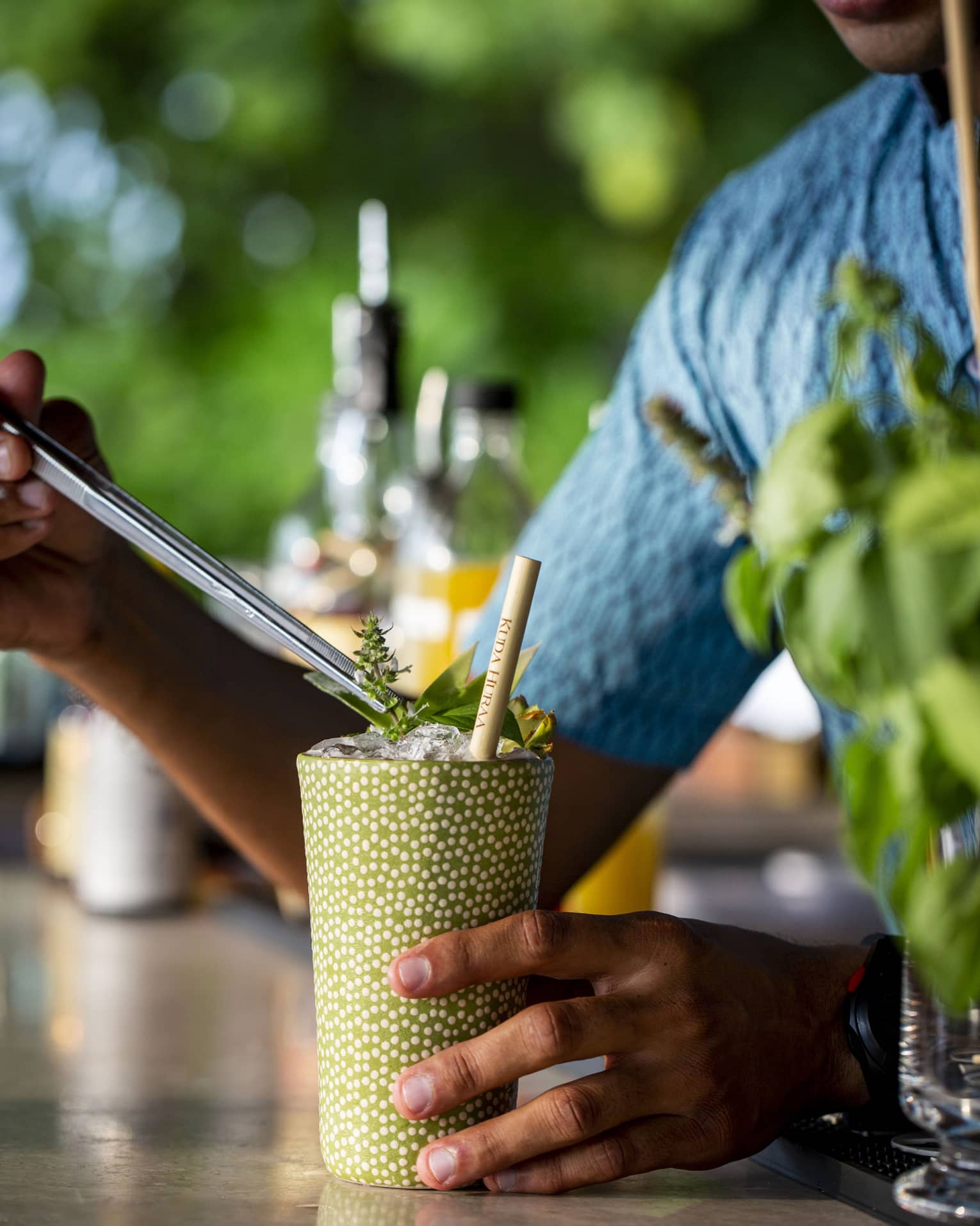 A smiling bartender in a blue shirt garnishes a cocktail; blurred bottles of liquor and green foliage in the background.