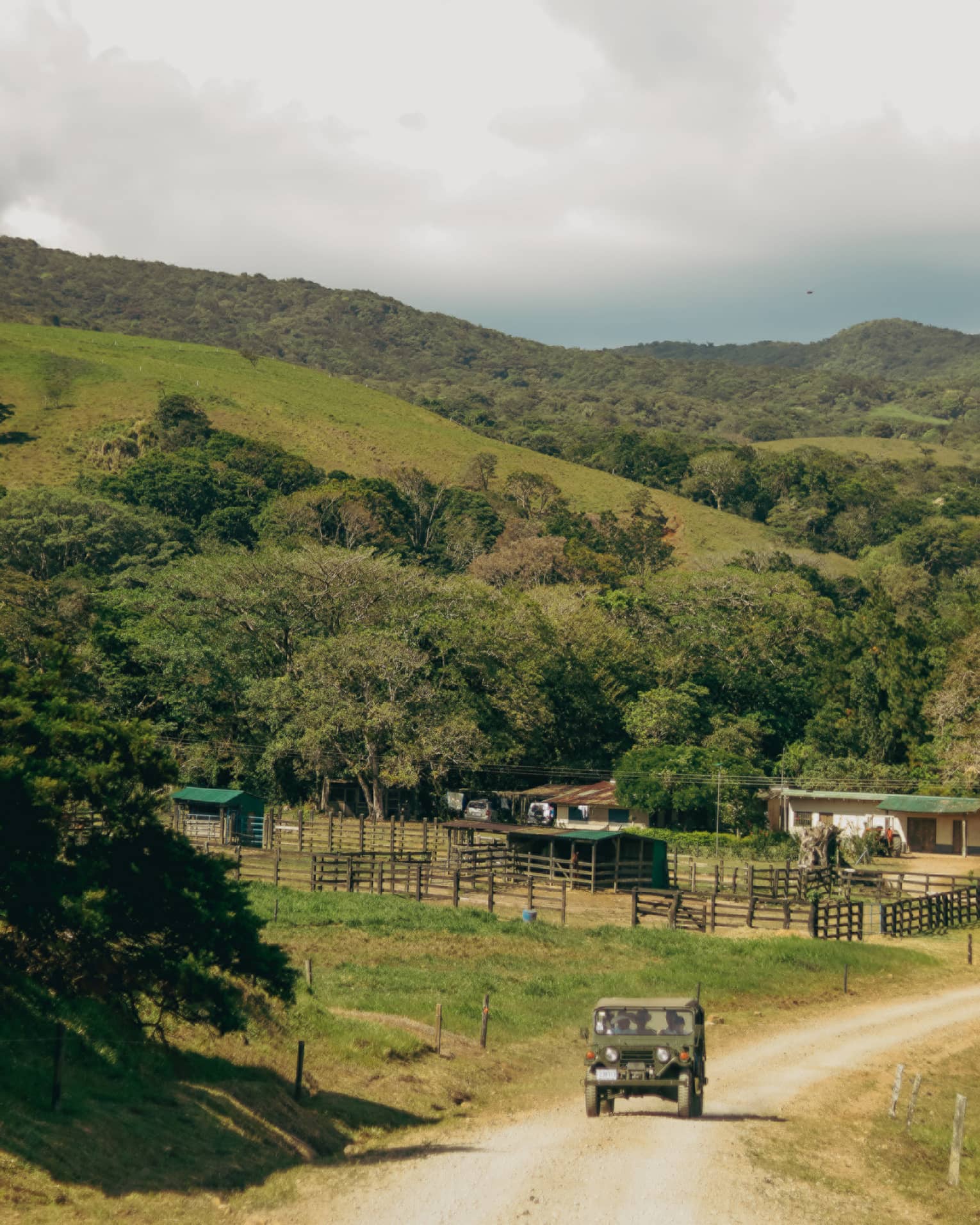 A vintage-style jeep rides along a dirt road, passing by a small farm in the hilly Costa Rican countryside