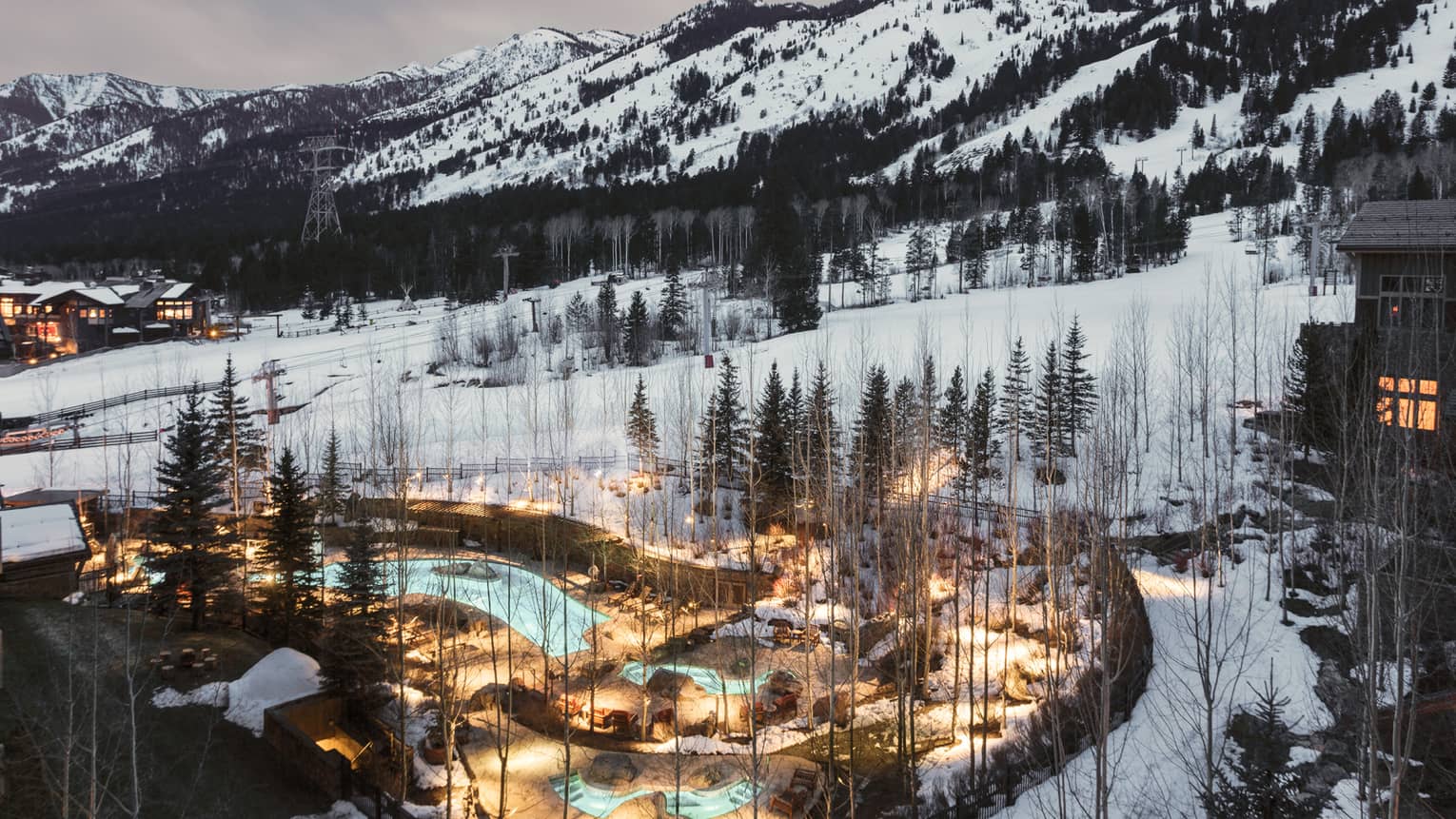 Aerial view of illuminated blue outdoor swimming pool through trees on snowy hill at dusk