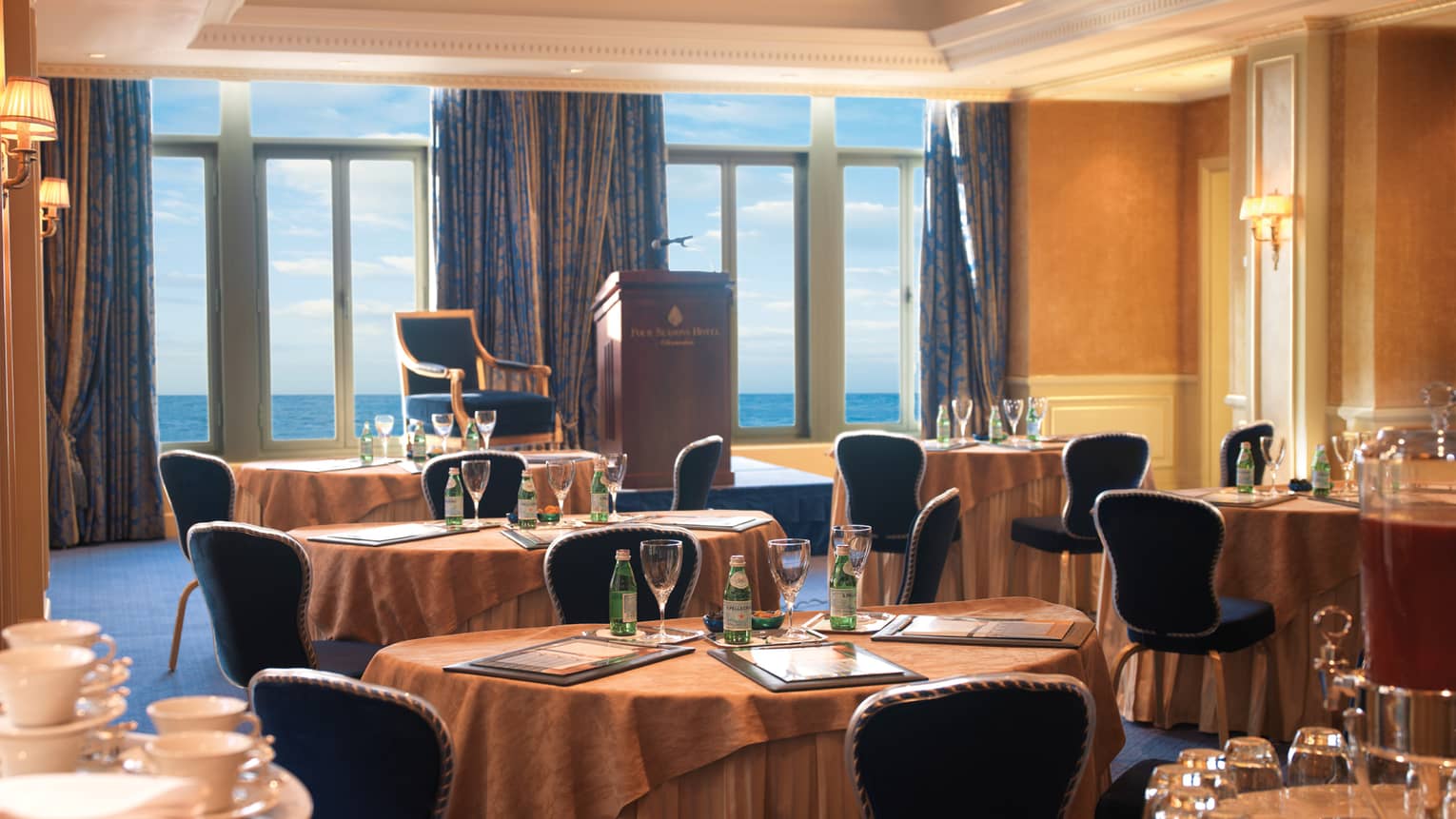 Sun-lit meeting room with round tables, royal blue chairs, podium facing floor-to-ceiling window and ocean views 