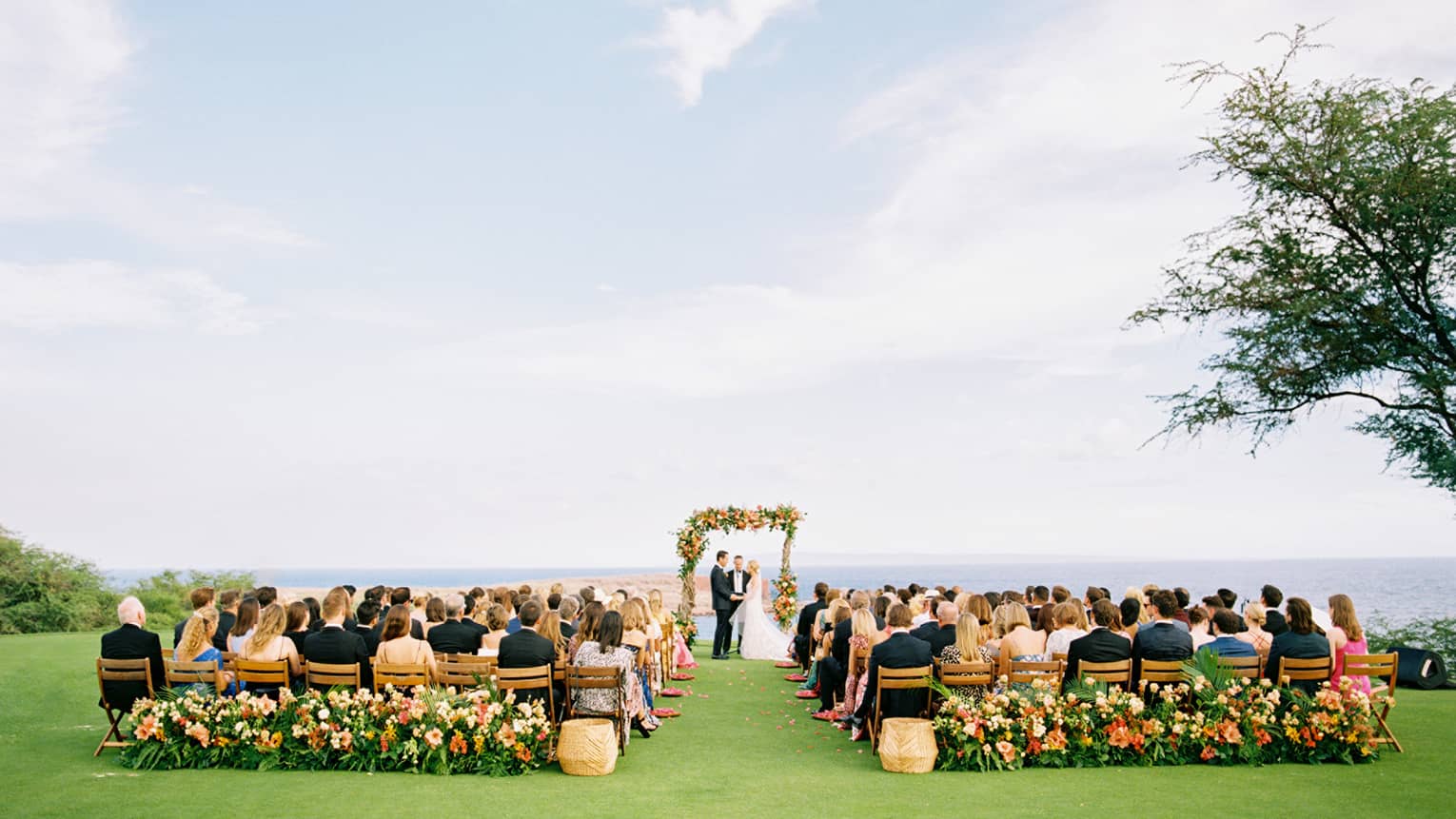 A wedding ceremony outside on a bright day, the bridge and groom are under a flower arch and many people are seated watching.