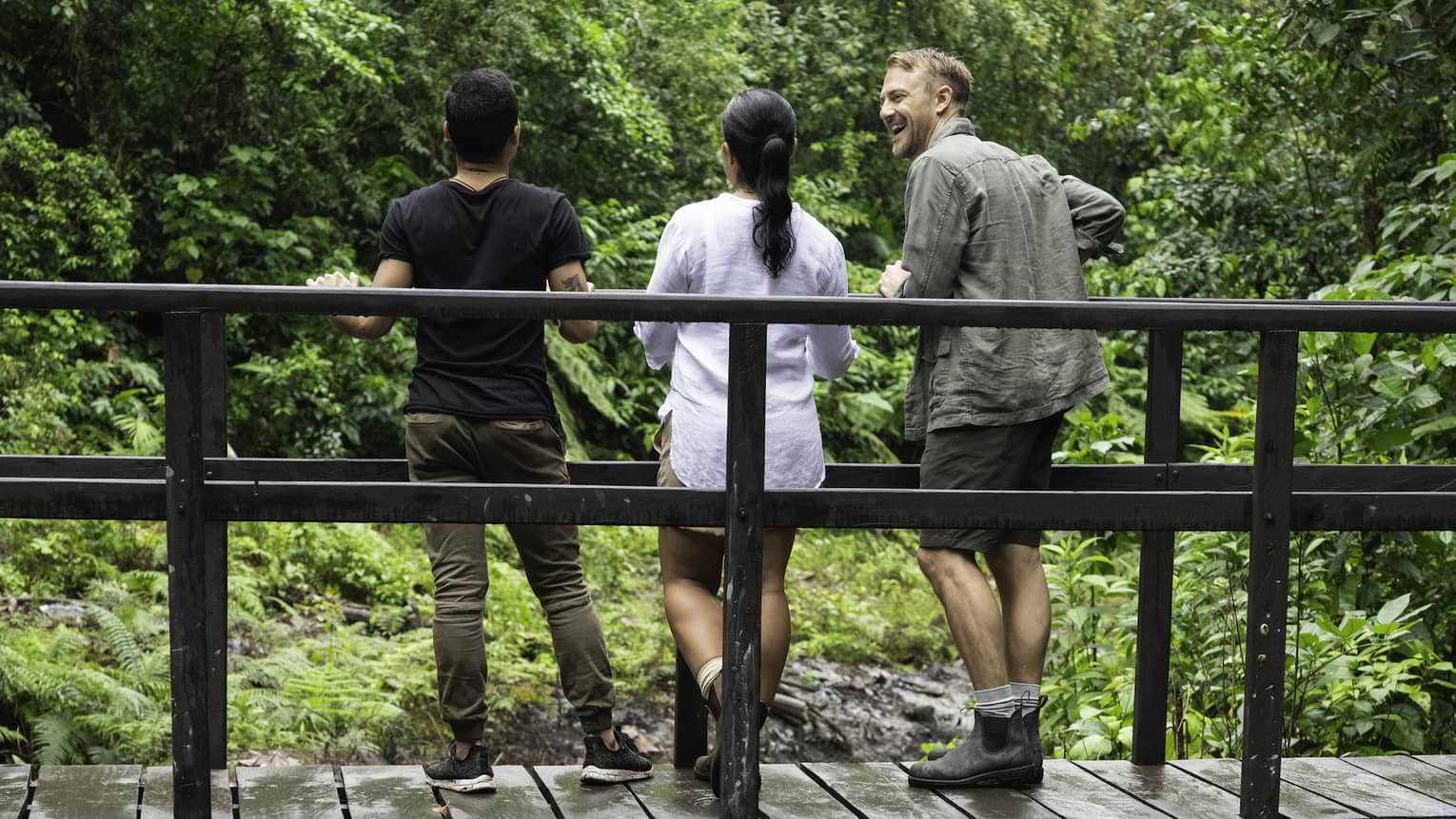 Three people stand on a wooden bridge that crosses over a small river in the middle of a forest