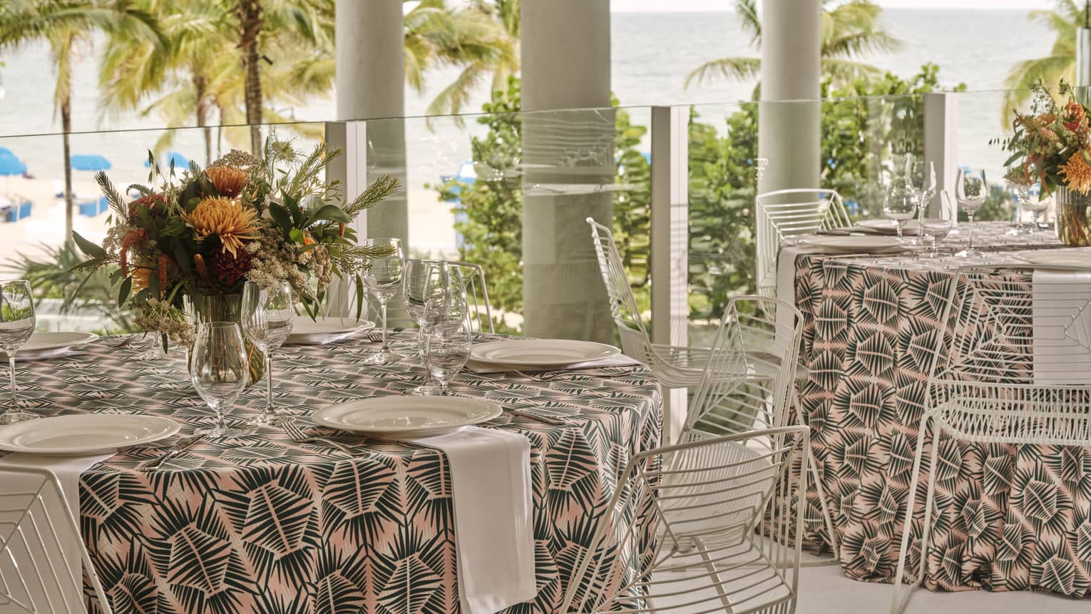 Round tables with table settings outside near a beach with palm trees.