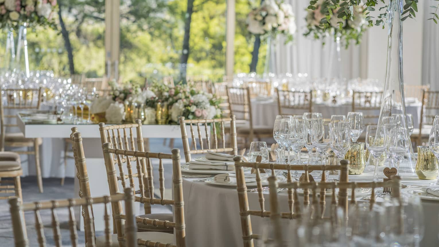 Round table with wooden chairs, place settings, glasses, floral display, in sunny room overlooking trees