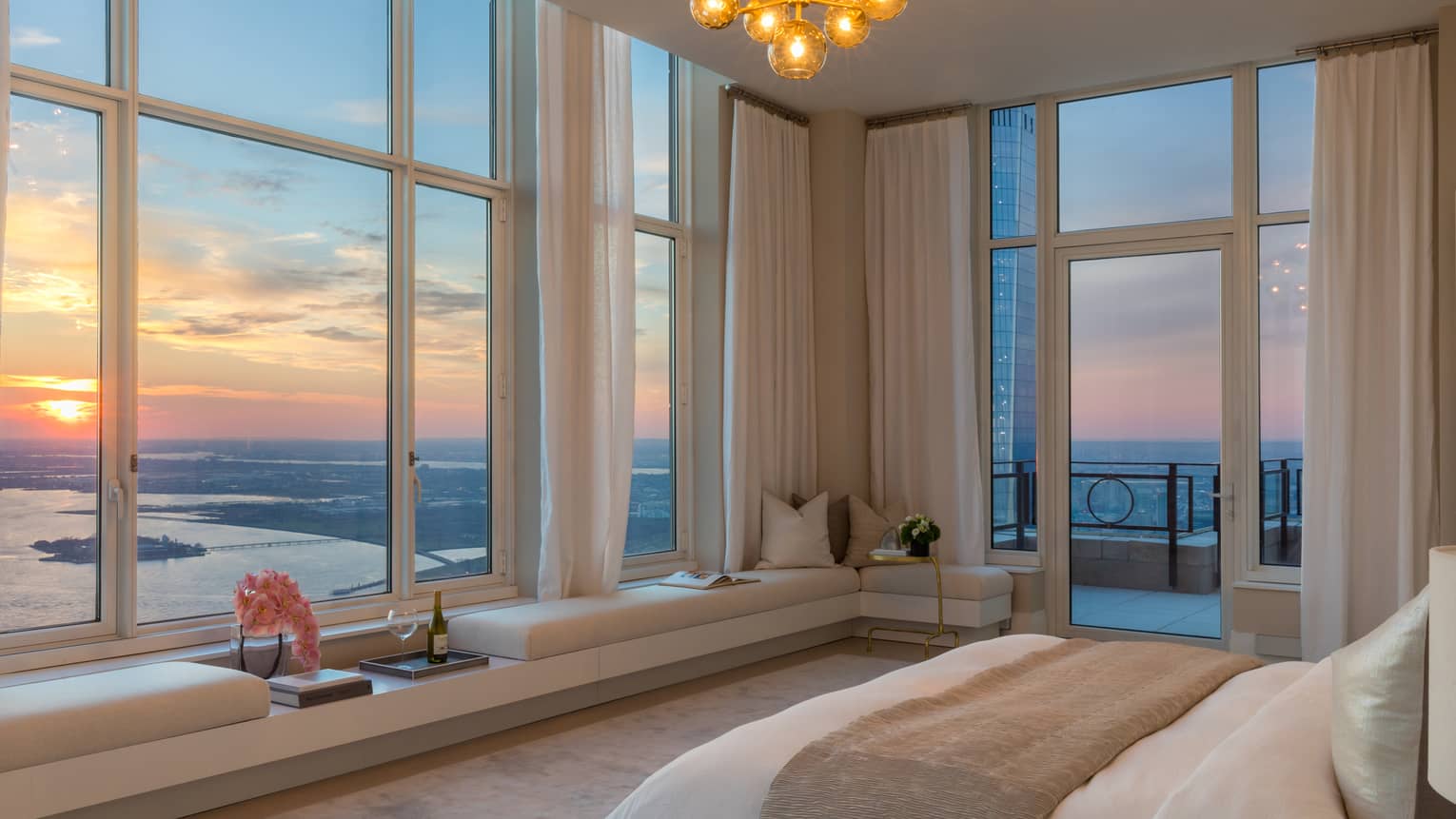 Bed with folded beige blanket across from corner picture windows, sunset views