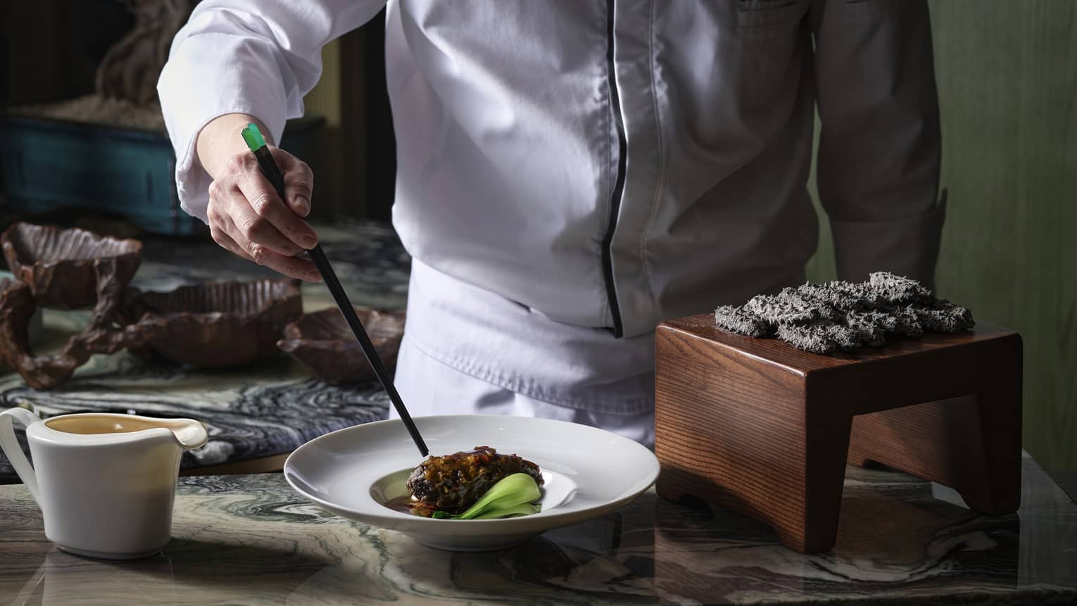 Chef using chopsticks to plate black pork belly in a white dish near a dozen black spiky delicacies on a small wood platform.