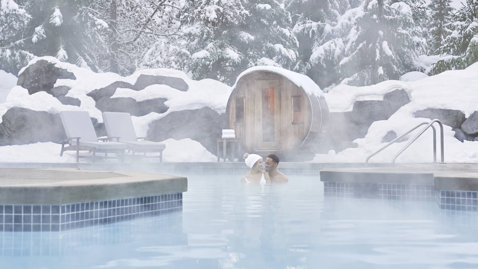 Couple wade in steaming outdoor swimming pool in winter, under snow covered rocks