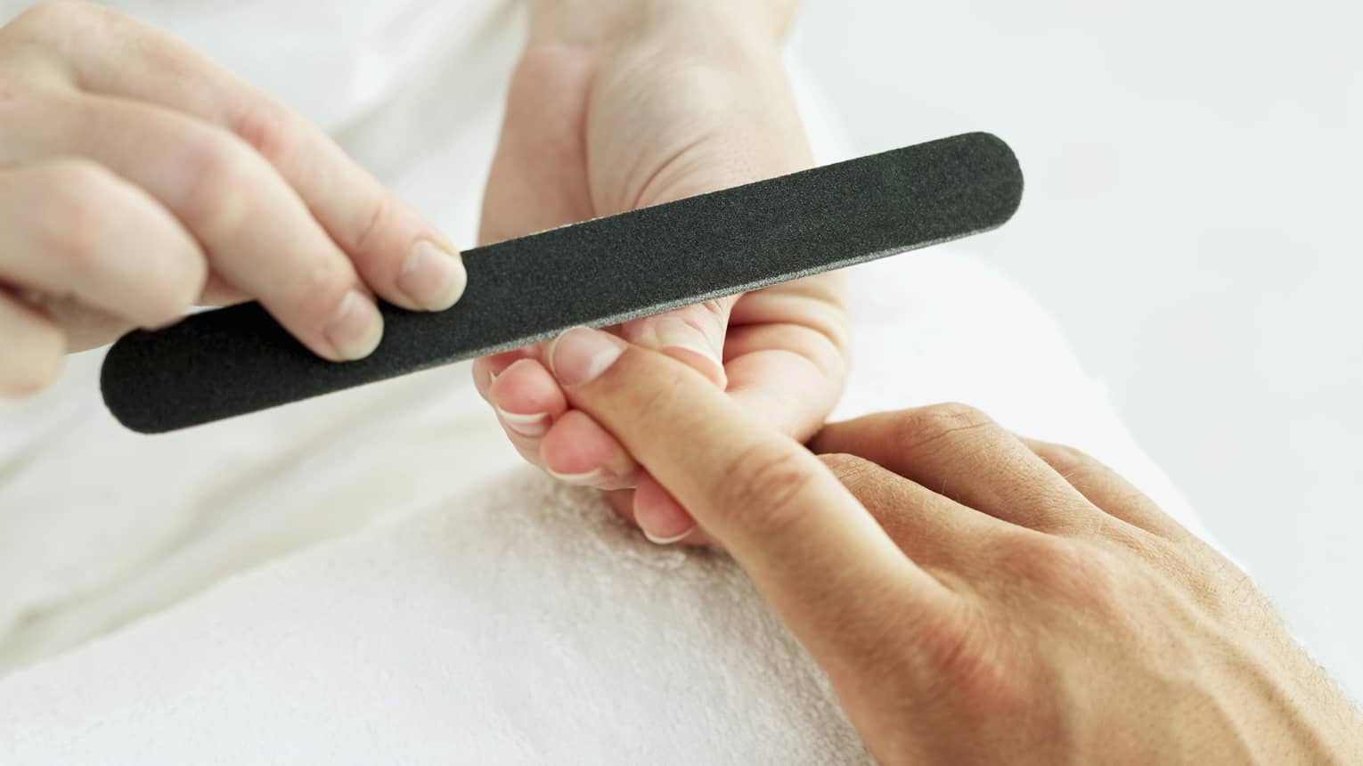 Woman holds manicure nail file to man's fingernails over white towel