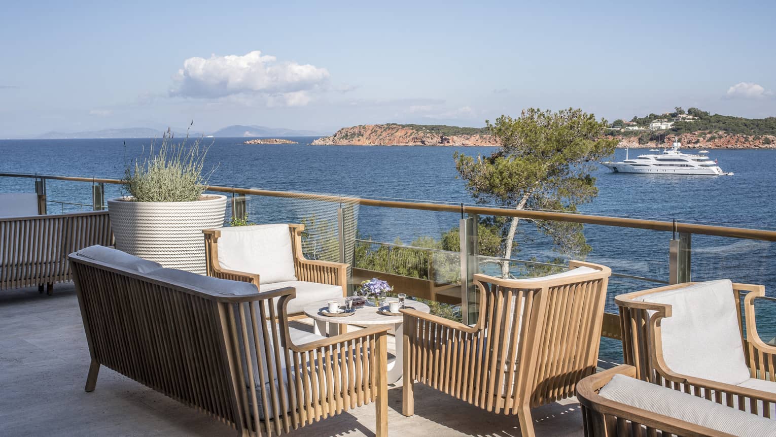 Wooden chairs with white cushions surrounding small round table by ocean with boat at Astron Terrace